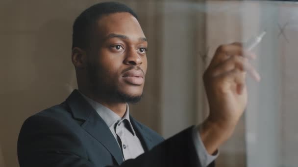 Portret afro amerykańskiego pracownika biznesu stojącego w biurze z markerem na przezroczystej szklanej ścianie robiącego notatki równanie rozwijające strategię projektu uwzględnia zysk finansowy uśmiechnięty — Wideo stockowe