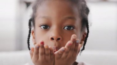 Yakın çekim mutlu yüz melez siyah çocuk kapalı mekanda, portre Afro Amerikan kızı küçük kız bebek afro çocuk kameraya bakıyor dostça bir yüz ifadesiyle hava öpücüğü, sevgi sembolü
