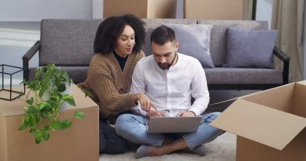 Casal multiétnico relaxar sentado no sofá usando laptop de computador para compras on-line fazer a ordem na loja de Internet em torno de caixas de papelão, menina afro-americana e homem caucasiano feliz movendo-se para nova casa — Vídeo de Stock