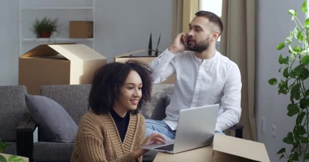 Casal multiétnico coletando coisas para se mudar para a nova casa chamando táxi usando smartphone, mulher afro-americana olhando para caixa de papelão vazia, homem caucasiano irritado chamando serviço de entrega de telefone celular — Vídeo de Stock