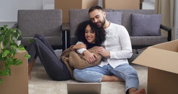 Jóvenes cansados afroamericanos rizado mujer y blanco caucásico hombre se sientan abrazando en el suelo cerca de cajas de cartón y plantas de interior resto después de mudarse a casa nueva apartamento, compra de bienes raíces — Vídeo de stock