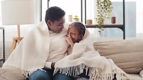 Afro american single mother parent mom woman hugs little black ethnic african girls child daughter sister, family talking secret sitting on couch covered with blanket, baby female kid shows one finger — Stock Video