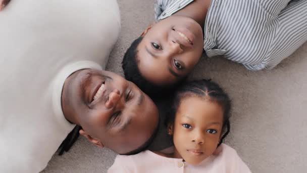 Портрет афроамериканських батьків з маленькою донькою лежить на підлозі, дивлячись на усміхненого дорослого батька-афроамериканця, який вказує на камеру пальцем, вгорі, на три покоління. — стокове відео