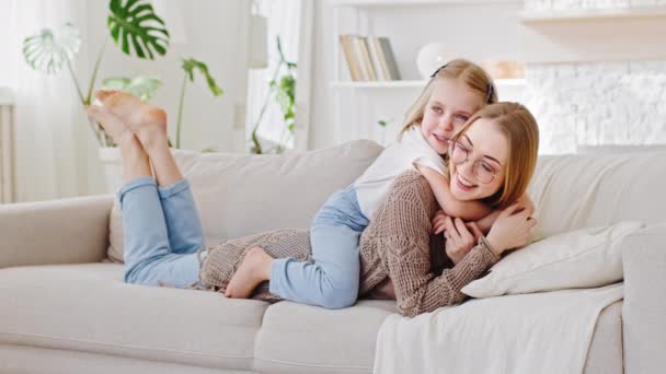 Bebê com a mãe descansando interior no sofá na acolhedora sala de estar rindo falando no fim de semana em casa, filha pequena menina abraços abraços abraços jovem mãe no pescoço sentado piggyback na mãe de volta, ligação familiar — Vídeo de Stock