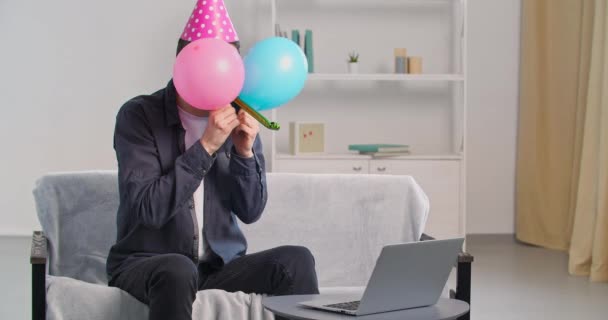 Glada rolig man bär födelsedag rosa hatt mössa ha kul ensam hemma tittar i laptop webkamera firar semester fest på distans med Internet vänner gömmer sig bakom ballonger blåser visselpipa — Stockvideo