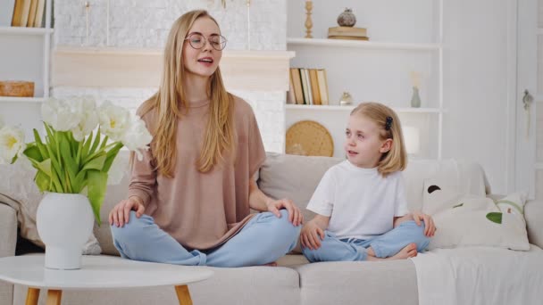 Young mother mindful mum teaches child beloved little daughter baby meditate sitting in lotus position on couch together, kid saying talking asking mother tries do yoga asana exercise, home practice — Stock Video
