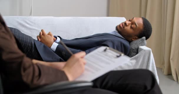 Homem de negócios afro-americano triste no terno elegante encontra-se no sofá no escritório do psiquiatra fala sobre problemas de emoções notícias tristes negativas psicólogo irreconhecível escreve a queixa de pacientes — Vídeo de Stock