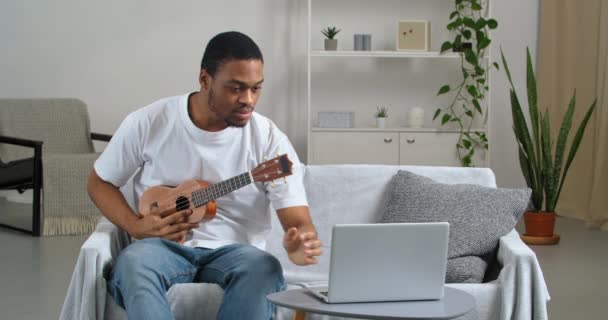 Συγκεντρωμένος Αφρο-Αμερικανός άντρας φοράει καθημερινά ρούχα βλέποντας video lesson παίζοντας κιθάρα σε απευθείας σύνδεση laptop επαναλαμβάνοντας πάνω από το διαδίκτυο δάσκαλος σπουδάζει εξάσκηση με μουσικό όργανο ukulele — Αρχείο Βίντεο