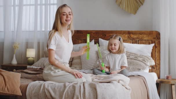 Caucasian family having fun at home in bed, single mother young adult woman mom with little daughter preschool girl child toddler baby kid in bedroom in morning blowing soap bubbles playing together — Stock Video