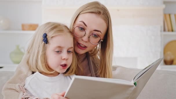 Portrait of two people family young single mother blonde woman mum wearing glasses caring mom reads literary book fairy tale to little daughter child toddler preschool kid sitting at home on weekend — Stock Video