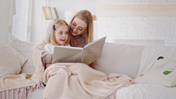 Inteligente madre adulta niñera lectura libro literatura cuento de hadas a pequeña hija colegiala preescolar niño sentado juntos cubierto con manta en casa en el sofá discutir hablar riendo — Vídeo de stock