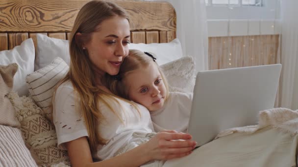 Jovem mãe solteira mãe adulta encontra-se na cama aconchegante com menina filha criança de manhã na noite de dormir antes de dormir olhando para a tela do laptop assistindo filmes fazer videoconferência de chamadas de casa — Vídeo de Stock