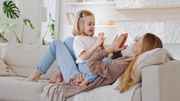 Cute little kid daughter with mom lying on sofa playing patty cake clap hands give five with palms in home, happy single mother and small child preschool girl enjoying funny game having fun on sofa — Wideo stockowe