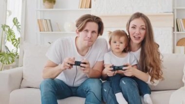 Aşırı neşeli beyaz anne-baba, baba ve küçük tatlı kızı olan anne bebek oyunu kazananlar, kanepe üzerinde video oyunu oynayanlar, anne ve kız çocuğuyla eğlenen heyecanlı baba video oyunu konsolları.