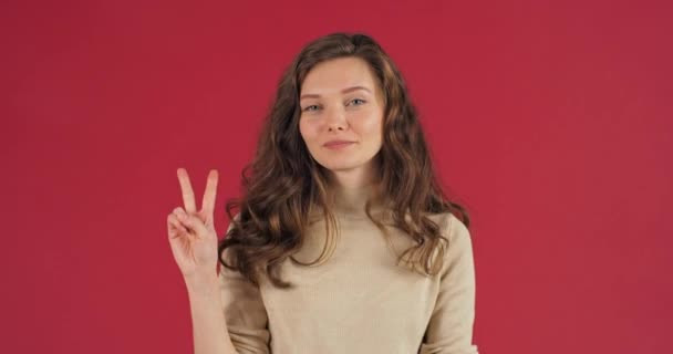 Positieve Kaukasische jonge meisje vrouw poseren op rode achtergrond in de studio tonen vrede gebaar twee vingers symbool van de overwinning vriendschap groeten, demonstreren v teken met de handen, lichaamstaal — Stockvideo