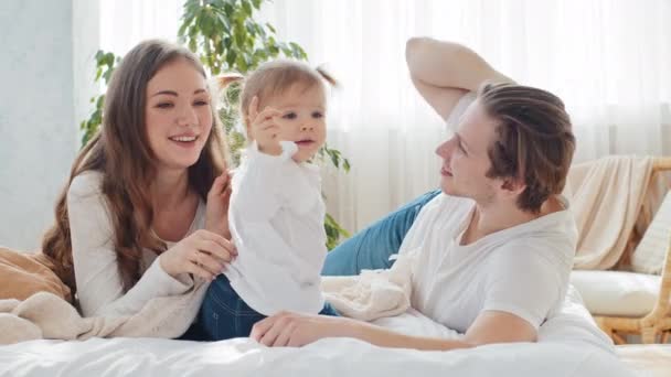 Caucasian young parents happy mother and caring father lie on bed with little girl child toddler daughter infant newborn chatting talk having fun enjoy family bonding love, kid points finger to camera — Stock Video