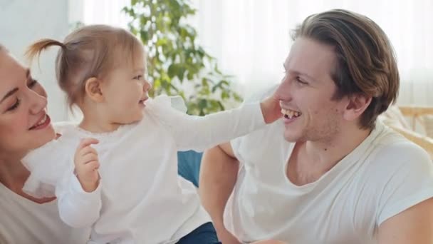 Young loving parents mom mother and happy father hug cuddle embrace little girl daughter toddler baby infant kiss beloved adorable kid on cheek lie together on bed at home enjoy family relationship — Stock Video