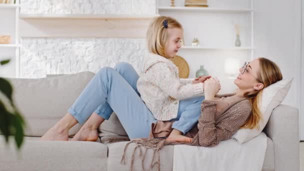 Happy mother woman babysitter with little cute girl daughter lie on couch relax rest at weekend talking chatting communicating together holding hands, mum speaks to kid child baby lying on home sofa — Stock Video