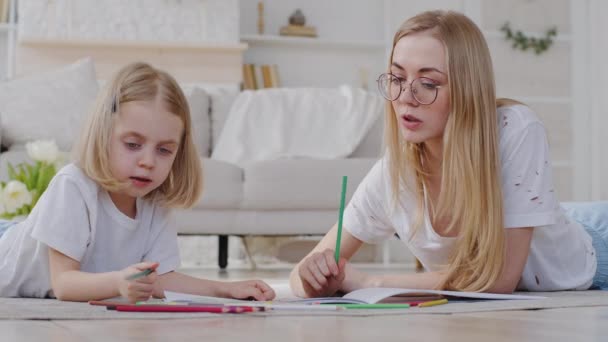 Αρχική εκπαίδευση, μαμά με κόρη κοριτσάκι μαθαίνουν να ζωγραφίζουν ξαπλωμένη στο πάτωμα του σαλονιού, δημιουργική δασκάλα μητέρα βοηθώντας το παιδί προσχολικής ηλικίας με το σπίτι ζωγραφίζοντας εικόνα με μολύβια, έννοια χόμπι — Αρχείο Βίντεο