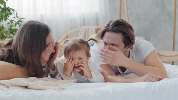 Happy caucasian parents caring mom and loving father playing hide and seek with toddler baby infant child kid daughter girl lying on bed in cozy home, family closes eyes with hands enjoy game together — Stock Video