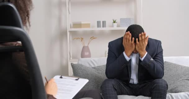 Afro-americano homem de negócios cara senta-se no sofá fala sobre seus problemas sente estresse emoções ruins gestos com as mãos segurando a cabeça de tristeza enquanto mulher psicólogo consulta escreve notas — Vídeo de Stock