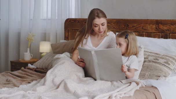 Caucasian young adult single mother woman mom reading fairy tale book to beloved little daughter girl child toddler kid baby at night before sleeping family lies in bed in cozy bedroom at home evening — Stock Video