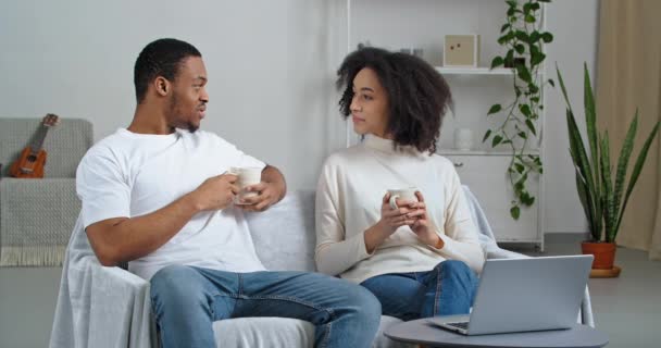 Pareja afroamericana familia jóvenes marido y mujer sentado en el sofá en acogedora sala de estar beber café de té caliente cacao disfrutando de fin de semana juntos discutir qué película para ver en línea en el ordenador portátil — Vídeos de Stock