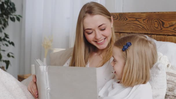 Portrait mom and daughter lie in bed resting hugging in bedroom at home, mother reads book at night before sleeping bedtime to little girl to child tells fairy tale story talking with baby kid smiling — Stock Video