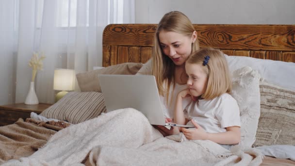 Young adult woman mom Caucasian mother hugs cuddles little daughter child baby look together in laptop screen choose movie online shopping in net make video call talking smiling lying in family bed — Stock Video