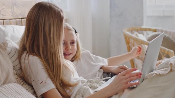 Mamá y su hija yacen en la cama mirando en el portátil. Madre soltera madre padre abrazos niña niño niño pequeño bebé utiliza la computadora para compras en línea navegación por Internet viendo película relajante en el dormitorio — Vídeo de stock