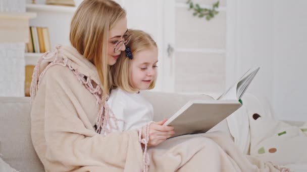 Giovane madre caucasica premurosa mamma donna bionda siede con la figlia sul divano lettura libro letteratura fiaba insegna bambino piccolo bambino bambina bambina letto coperto con coperta riposo sul divano di casa — Video Stock