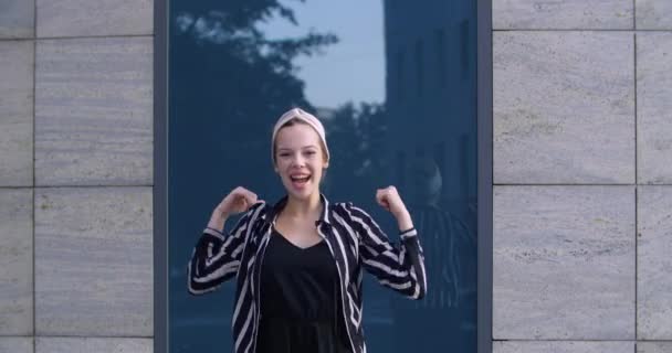 Jong meisje student draagt stijlvolle hoofdband en gestreepte jas staat alleen buiten op straat schreeuwt luid van geluk opent haar mond vieren overwinning triomf komisch beweegt armen grappige dansen — Stockvideo
