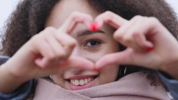 Vonzó barátságos Afro amerikai lány imádott nő mutatja szív gesztus ujjaival néz kamera és mosolyog, vegyes faji hölgy göndör buja haj pózol mutatja jelképe kedvesség Valentin-napon — Stock videók