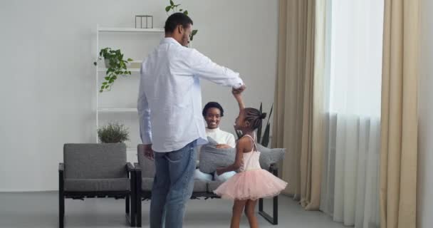 Älskar afro amerikansk pappa håller hand med sin dotter dansar med söta små barn prinsessa bär rosa klänning i modernt vardagsrum, lycklig far och liten flicka spelar show ung mor dans — Stockvideo