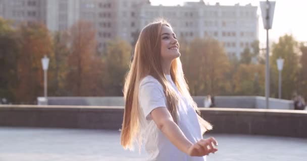 Egy fiatal, gyönyörű, szőke diák alkalmi ruhában, örömtánccal, felemelt kezekkel a város központjában, napfényben. Gyönyörű lány vonzó divat női modell repülő haj — Stock videók