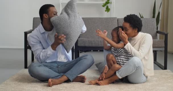 Afro american family sitting on floor at home on weekend, young adult african mother hugging little black girl child kid, daughter showing tongue to dad, father playing with pillow laughing talking — Stock Video