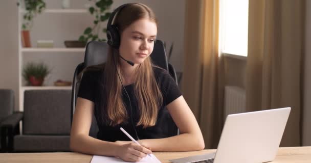 Дружелюбная кавказская старшеклассница носит наушники и концентрируется на работе, глядя на экран ноутбука, общается с преподавателем онлайн, делает заметки, дистанционное обучение — стоковое видео