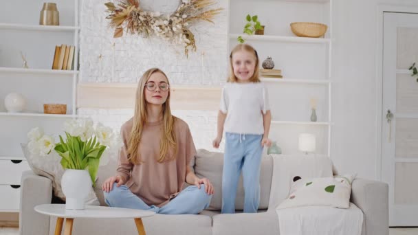 Bullriga barn aktiv liten flicka dotter unge hoppar på soffa baby dåligt uppförande distraherande störande ung mor lugn kvinna mamma meditera sitter i lotus position i vardagsrummet, lugn koncept — Stockvideo