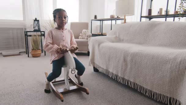 Afro-Amerikan kız çocuğu küçük çocuk anaokulu çocuğu oyuncak ata biniyor, evde yalnız başına eğleniyor iç mekanda oyun çocukluğunun tadını çıkarıyor, hafta sonları evde oynuyor. — Stok video