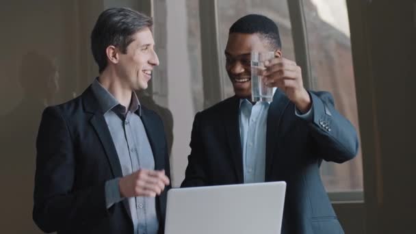 Dos hombres de negocios multirraciales arquitecto afroamericano e ingeniero inversor caucásico mostrando en portátil nuevo proyecto de construcción afro hombre levantando vaso de agua felicitando mirando por la ventana soñando — Vídeos de Stock