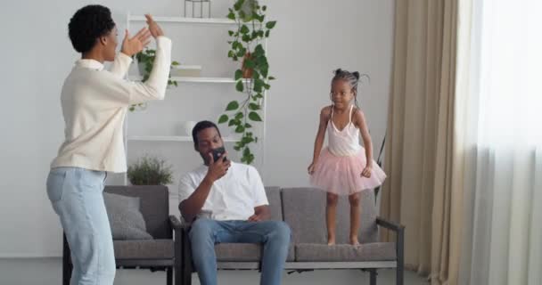 Afrikanische Familie verbringt Zeit zu Hause, schwarze Afro-Mutter gibt dem kleinen Kind im rosafarbenen Prinzessinnenkleid Befehl, von der Couch auf den weichen Teppich zu springen, der Vater sitzt lachend auf dem Sofa — Stockvideo