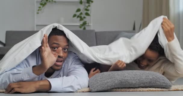 Afro american family hiding under warm white blanket appears waving hands hello shouting noisy smile laughing looking at camera, african father black mom and ethnic child have fun at weekend together — Stock Video