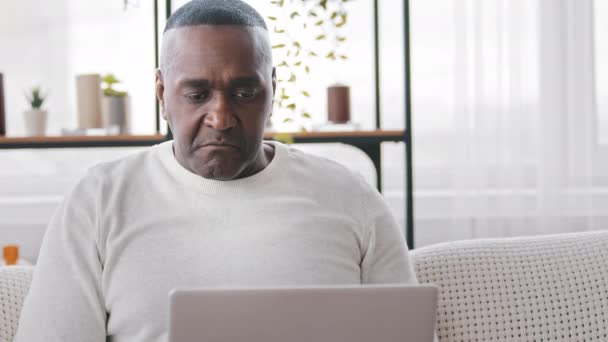 Skoncentrowany Afroamerykanin dojrzały w średnim wieku 50 lat dorosły mężczyzna czarny etniczny biznesmen siedzi na kanapie domu patrząc na laptopa czytanie wiadomości zakupy online w sieci pracy zdalnie za pomocą aplikacji komputerowej — Wideo stockowe
