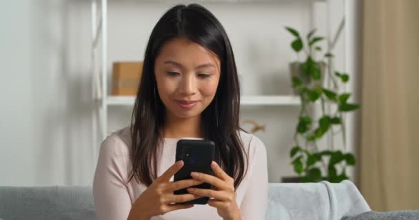 Retrato de asiático dona de casa mulher sentado no sofá em casa olhando para tela do telefone móvel jogando online com amigos recebe mensagem agradável sorrindo resposta de digitação navegador rolagem no aplicativo gadget — Vídeo de Stock