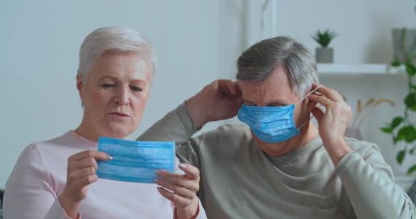 Idős pár, akik orvosi maszkot viselnek a régi arcukon, nagyapa és nagymama, akik gondoskodnak az egészségről védik a vírusfertőzéstől, elégedetlen 50-es évekbeli maszkos férfi feleséggel otthon — Stock videók