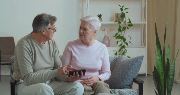 Ouderen paar oude vrouw en man kijken naar mobiele telefoon zitten op de bank thuis in de woonkamer ruzie over wat ze zagen op video online bespreken ontvangen SMS-bericht kwaad ruzie — Stockvideo