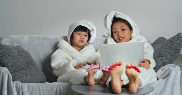 Mãe e filha relaxar em casa spa fazendo pedicure cuidar da pele assistindo filmes no laptop on-line vestindo roupões de banho brancos quentes, duas irmãs asiáticas de diferentes gerações fazendo tratamentos de beleza — Vídeo de Stock