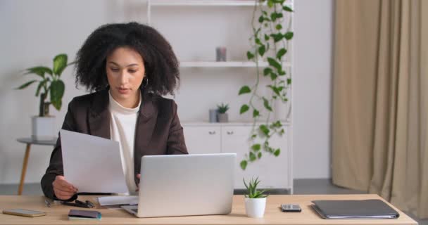 Афроамериканська студентка - секретар позаштатної ділової жінки, яка сидить за столом в офісі і дивиться на ноутбук, порівнює дані на папері і на екрані комп'ютера аналізує проект. — стокове відео