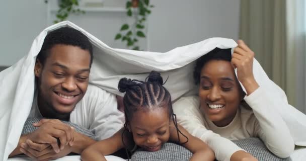 Keluarga Afro tergeletak di bawah selimut melihat ke kamera seperti jempol sampai berisik tertawa bersenang-senang bersama di rumah, ayah membuat isyarat persetujuan bayi berpura-pura tidur ibu menepuk pipi — Stok Video