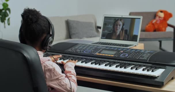 Menina irreconhecível afro-americana criança usa fones de ouvido faz chamada de vídeo para professor se comunica on-line com tutor aprender a tocar sintetizador de piano eletrônico, aprendendo durante bloqueio — Vídeo de Stock